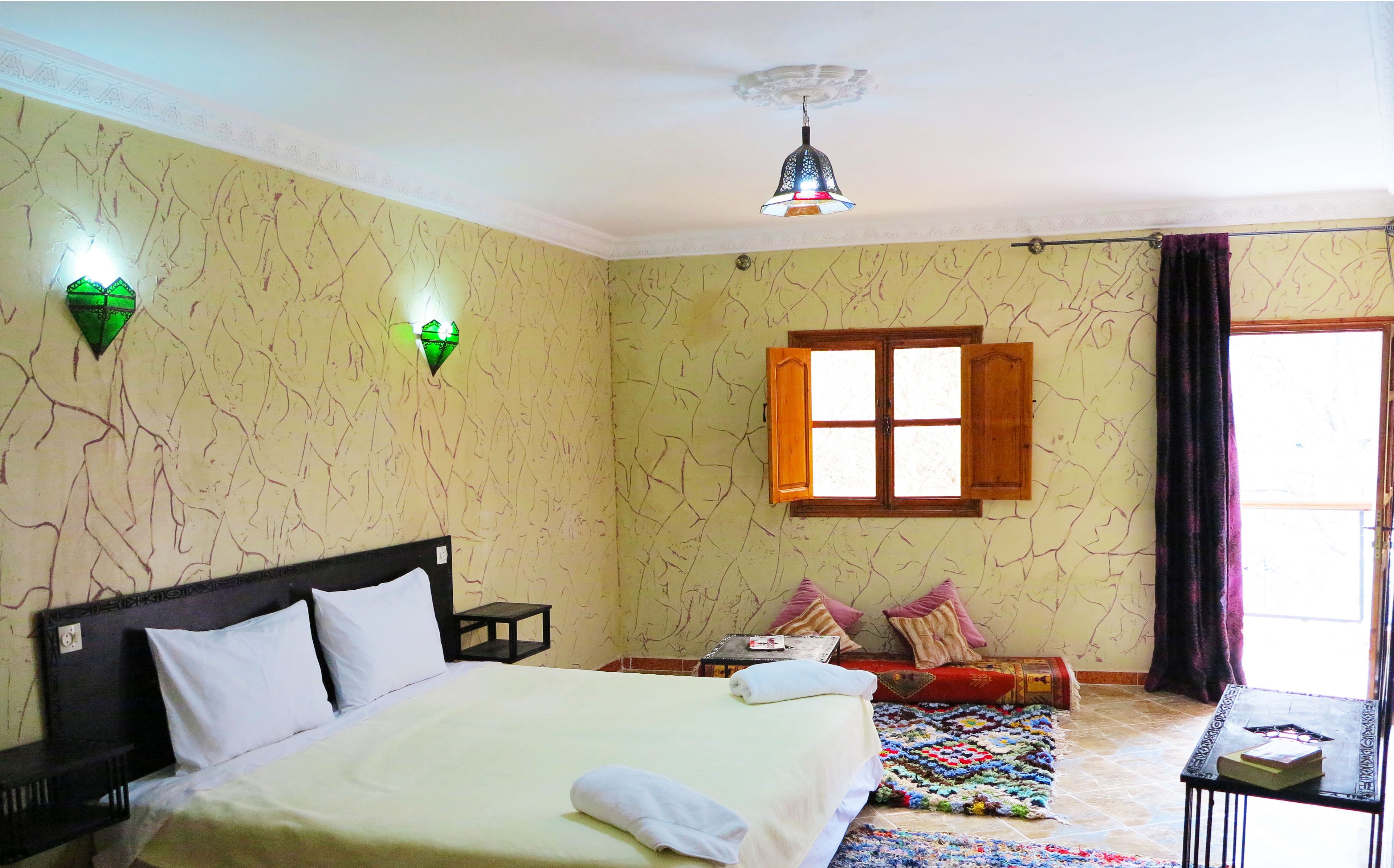 Habitación de hotel con una cama, una mesita pequeña, una ventana con cortinas y una lámpara de pared decorativa.