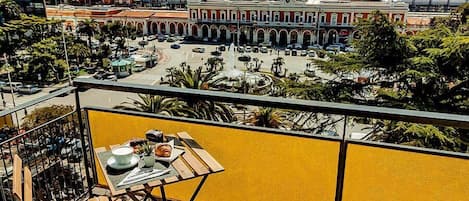 A balcony with a table set for two, overlooking a city square with buildings and a fountain.