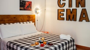 Quarto de hotel com colcha xadrez, bandeja de café da manhã com torradas, frutas e uma bebida, e uma placa na parede com a palavra 'CINEMA'.