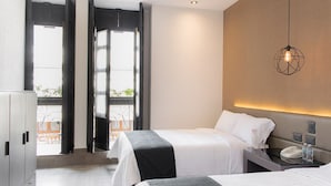 A hotel room with two beds, a bedside table with a phone and water bottle, a hanging light fixture, and a view of the outdoors through a large window.