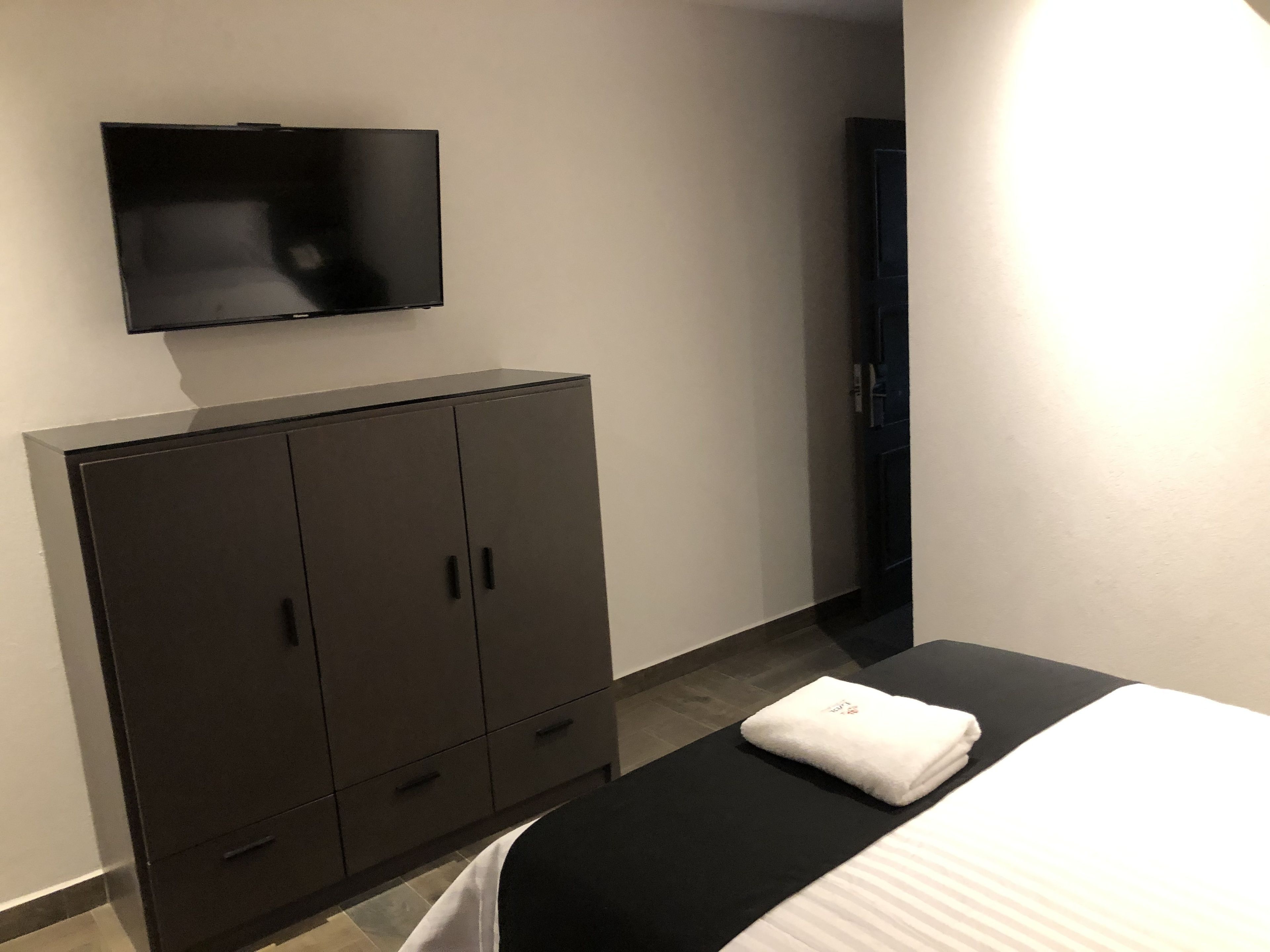 Habitación de hotel con cama, televisor montado en la pared, armario y una toalla sobre la cama.