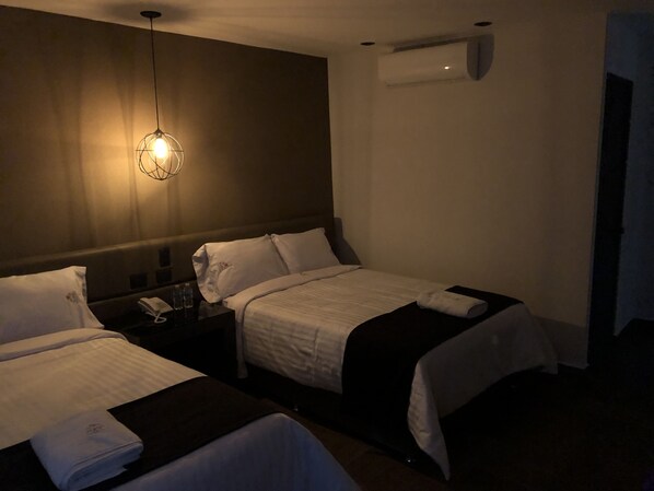 Une chambre d’hôtel avec deux lits, une table de chevet avec un téléphone et une bouteille d’eau, un luminaire suspendu et un appareil de climatisation fixé au mur.