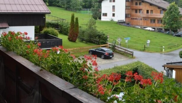 Une vue depuis un balcon donnant sur un bâtiment avec un toit rouge et une façade blanche, entouré de verdure et d’un paysage vallonné.