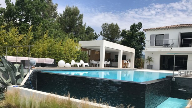 Un área de piscina moderna al aire libre con una piscina rectangular, rodeada por un muro de piedra y un porche cubierto con mobiliario blanco.