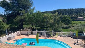 Piscina com borda curva, rodeada por uma cerca branca, e vista para uma paisagem montanhosa com árvores e edifícios ao fundo.