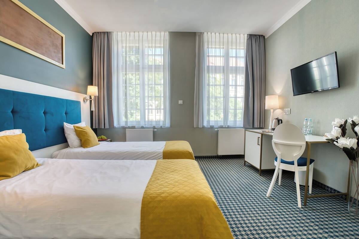 Habitación de hotel con dos camas, un escritorio, una silla, un televisor y ventanas amplias.