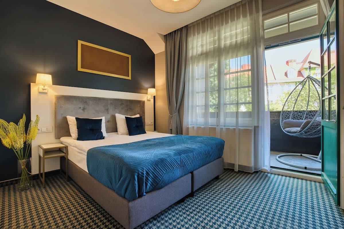 Un cuarto de hotel con cama, lámparas de noche, ventana con cortinas, un sillón colgante y vista al exterior.