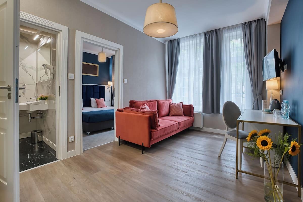 Una habitación de hotel moderna con un sofá rojo, una mesa de comedor con un jarrón de girasoles, un televisor y un baño con cabina de ducha de vidrio.
