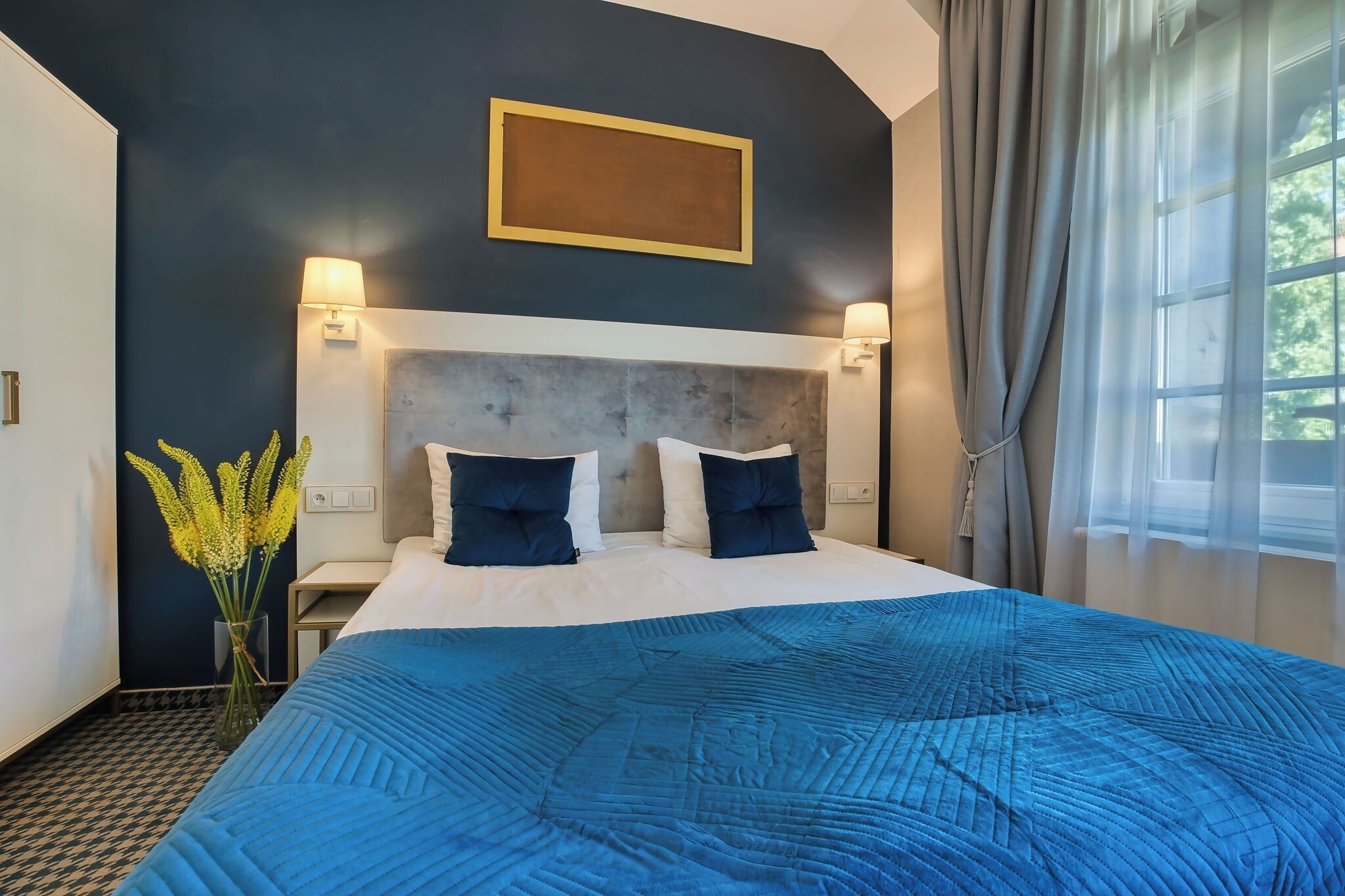 Un dormitorio con una cama grande, dos lámparas de pared, un cuadro enmarcado sobre la cama y una ventana con cortinas.