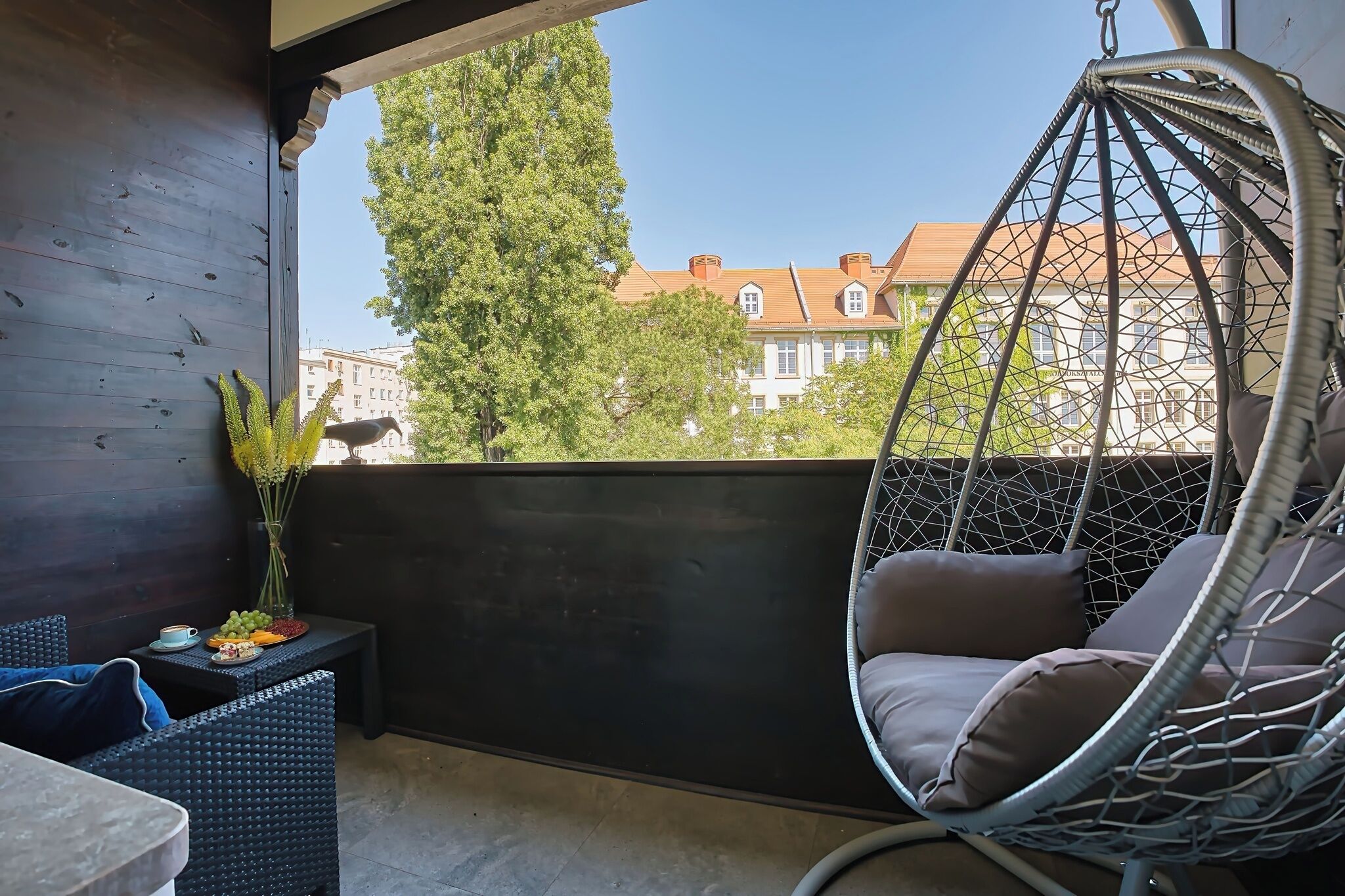 A balcony with a hanging chair, a small table with a vase and fruit, and a view of buildings and trees.