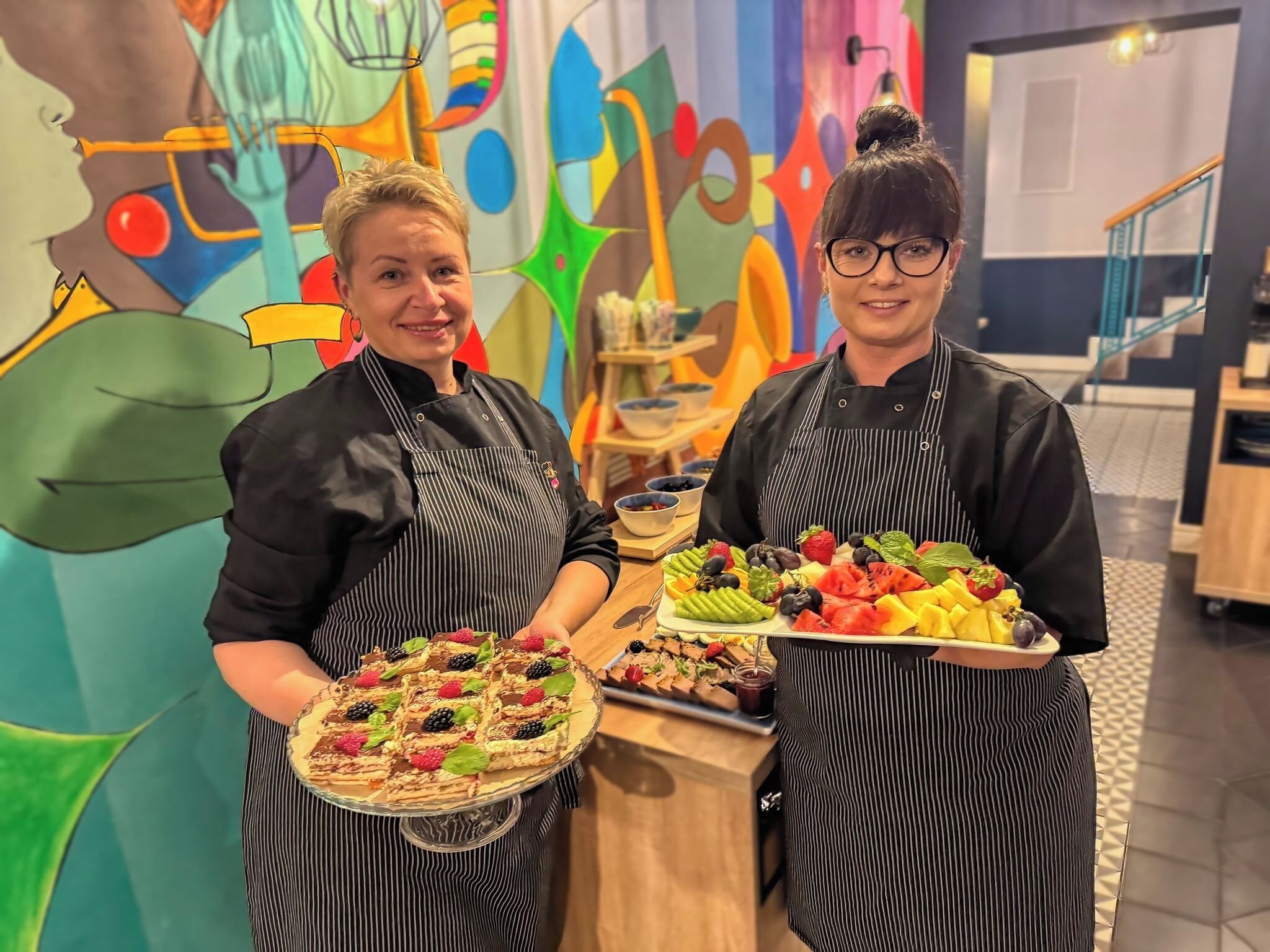 Two individuals in aprons holding trays of food, with a colorful abstract mural in the background.