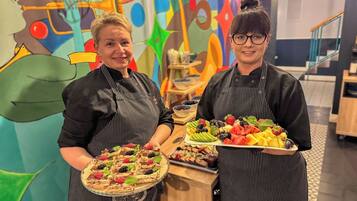 Two individuals in aprons holding trays of food, with a colorful abstract mural in the background.