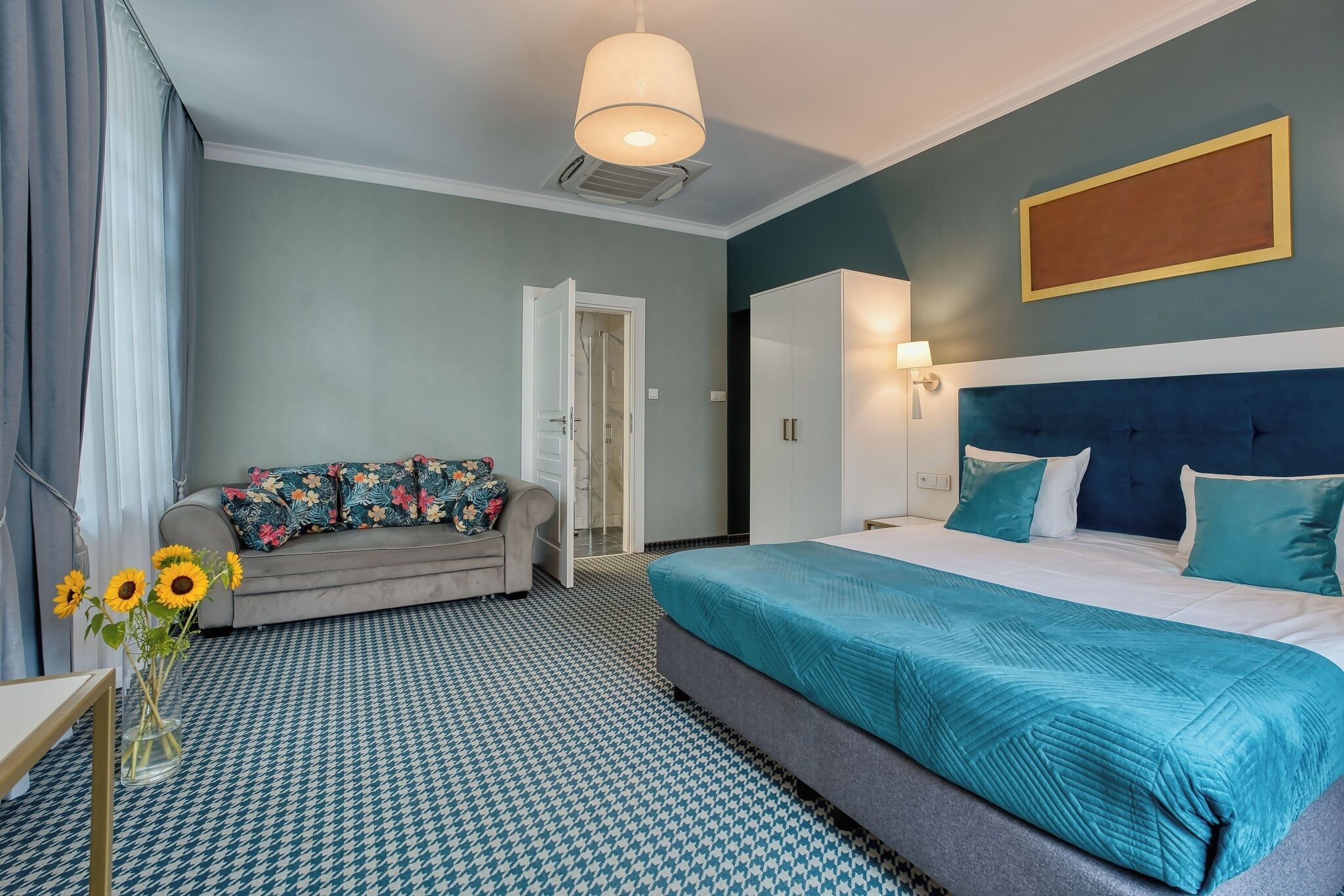 A hotel room with a bed, a sofa, a vase with flowers, and a patterned floor.