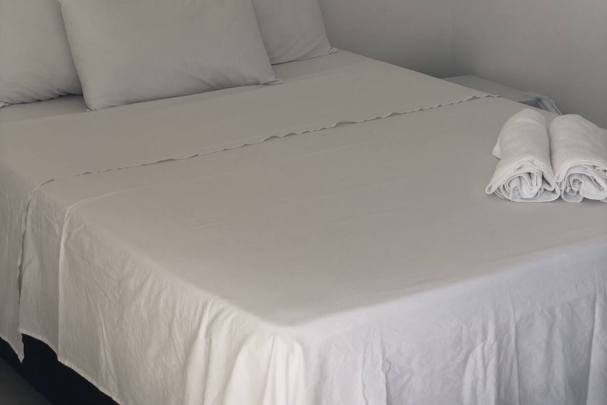 Un lit bien fait, avec du linge de lit et des oreillers blancs, une serviette pliée posée sur le pied de lit, et un mur sobre agrémenté d’un petit interrupteur.
