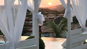 Ein weißes Gartenhaus mit transparenten Vorhängen und einem Handtuch, das am Gestell befestigt ist, mit Blick auf eine Wohngegend mit Häusern und Palmen bei Sonnenuntergang.
