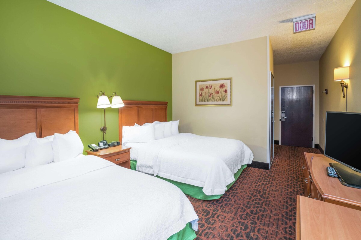 A hotel room with two beds, a desk with a TV, a lamp, and a painting on the wall.