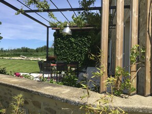 Um pátio externo com mesa e cadeiras, rodeado por vegetação e uma estrutura de pérgola.