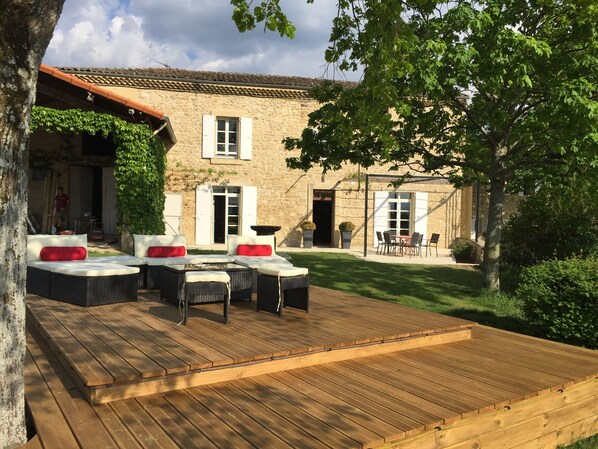 A stone building with a tiled roof, a wooden deck with outdoor furniture, and a well-maintained lawn.