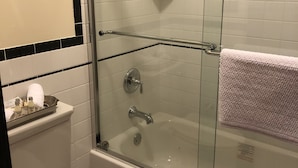 A bathroom with a glass-enclosed shower, a white toilet, and a glass shelf with toiletries. - Guest Quarters at the Inn  - 102 (North Beach)