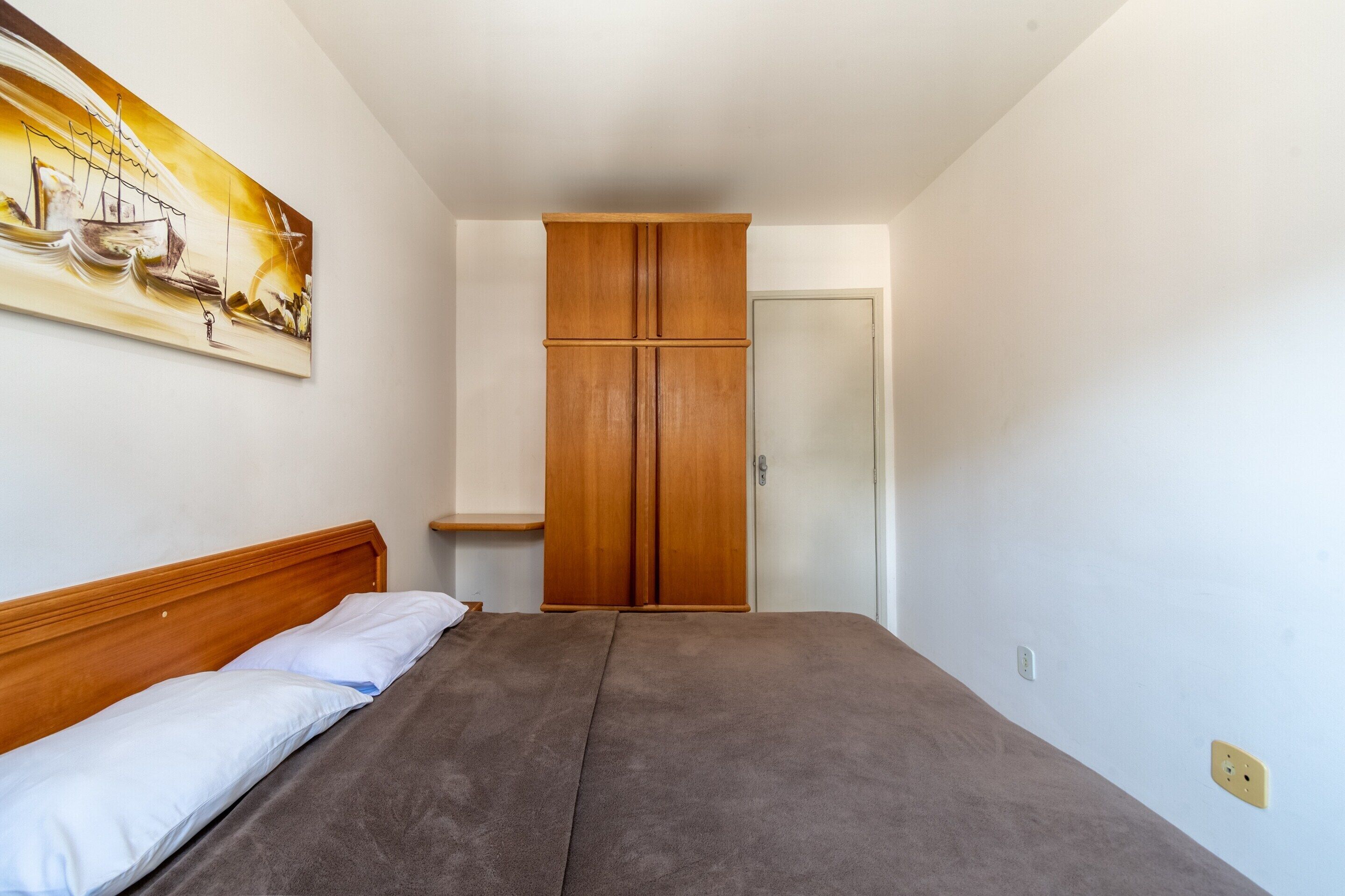 A bedroom with a bed, a wooden wardrobe, a painting on the wall, and a door.