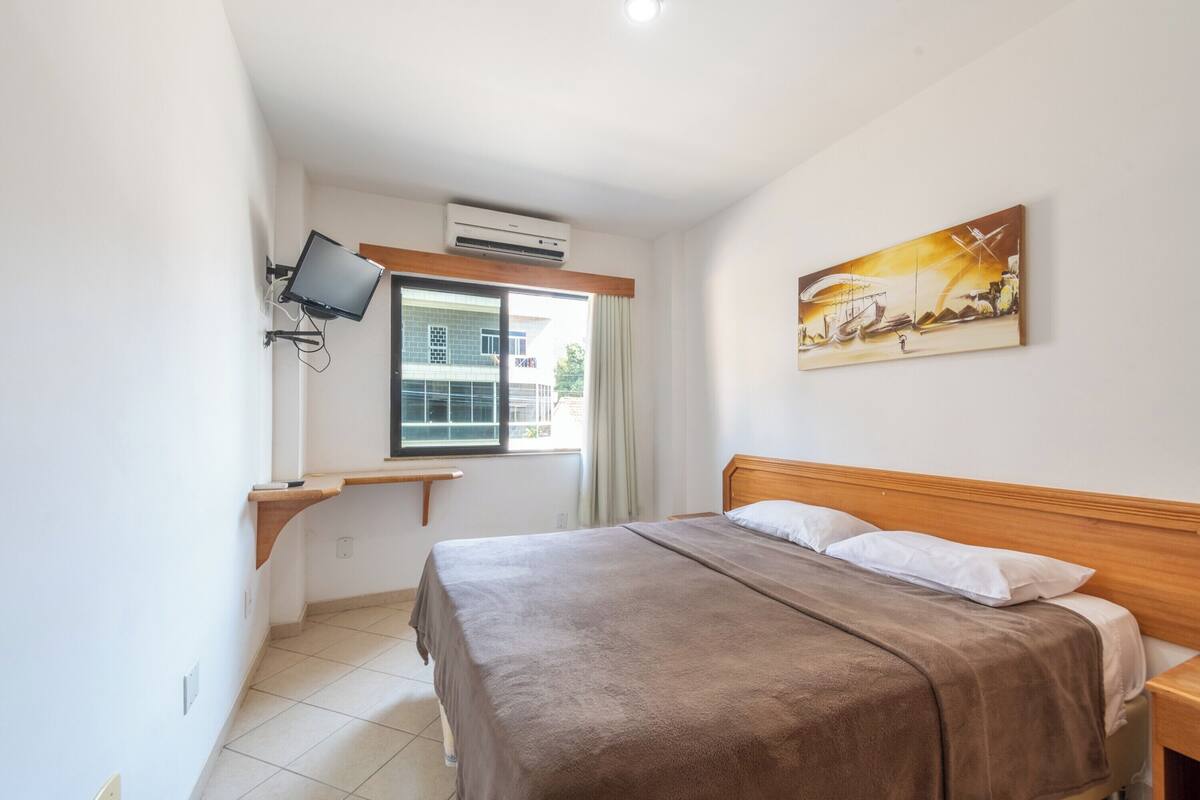 A bedroom with a bed, a TV mounted on the wall, a window, and a painting.
