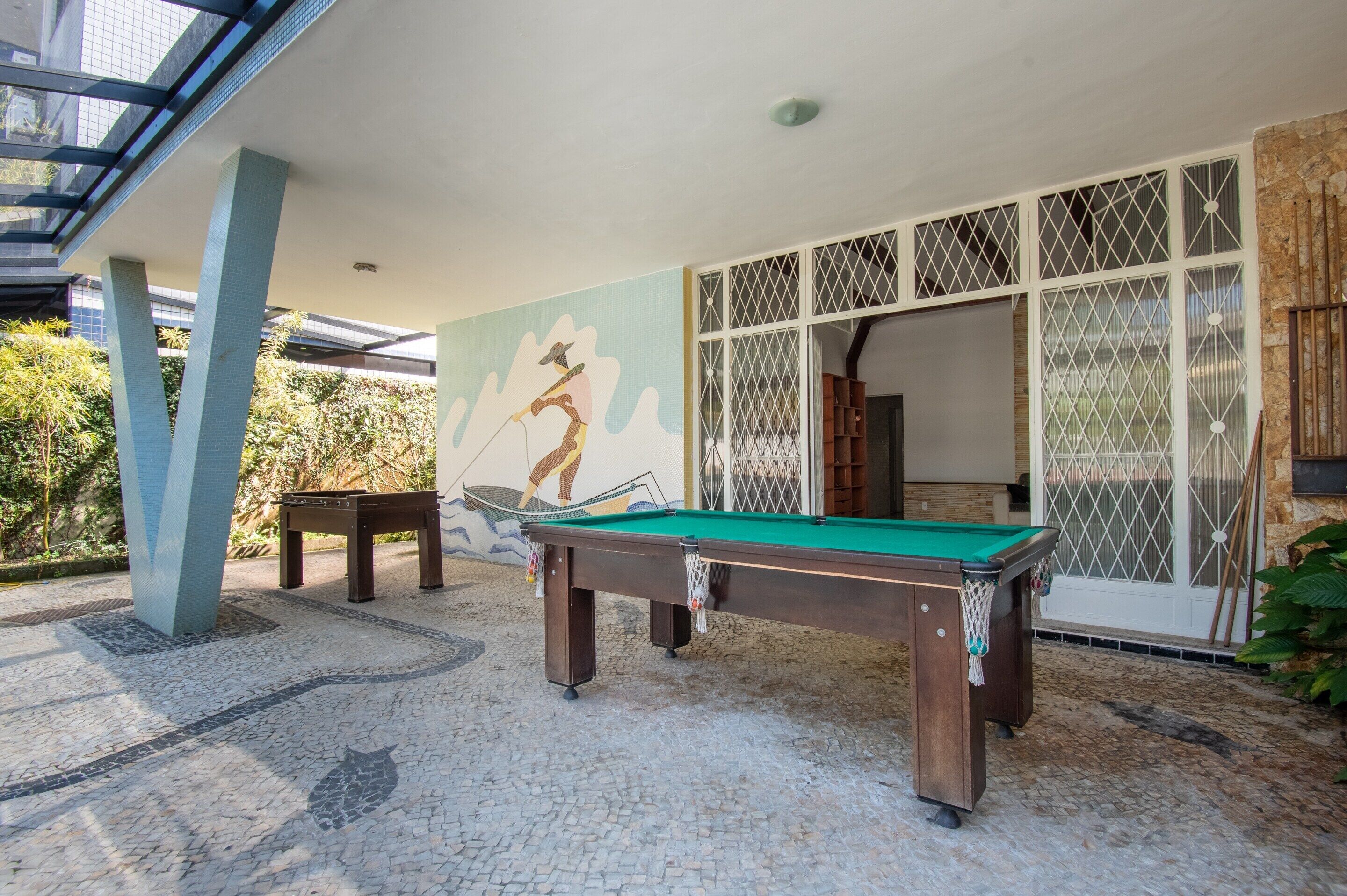 Uma mesa de bilhar ao ar livre com superfície em feltro verde, rodeada por um piso estampado, com um edifício de grandes janelas e um mural decorativo ao fundo.
