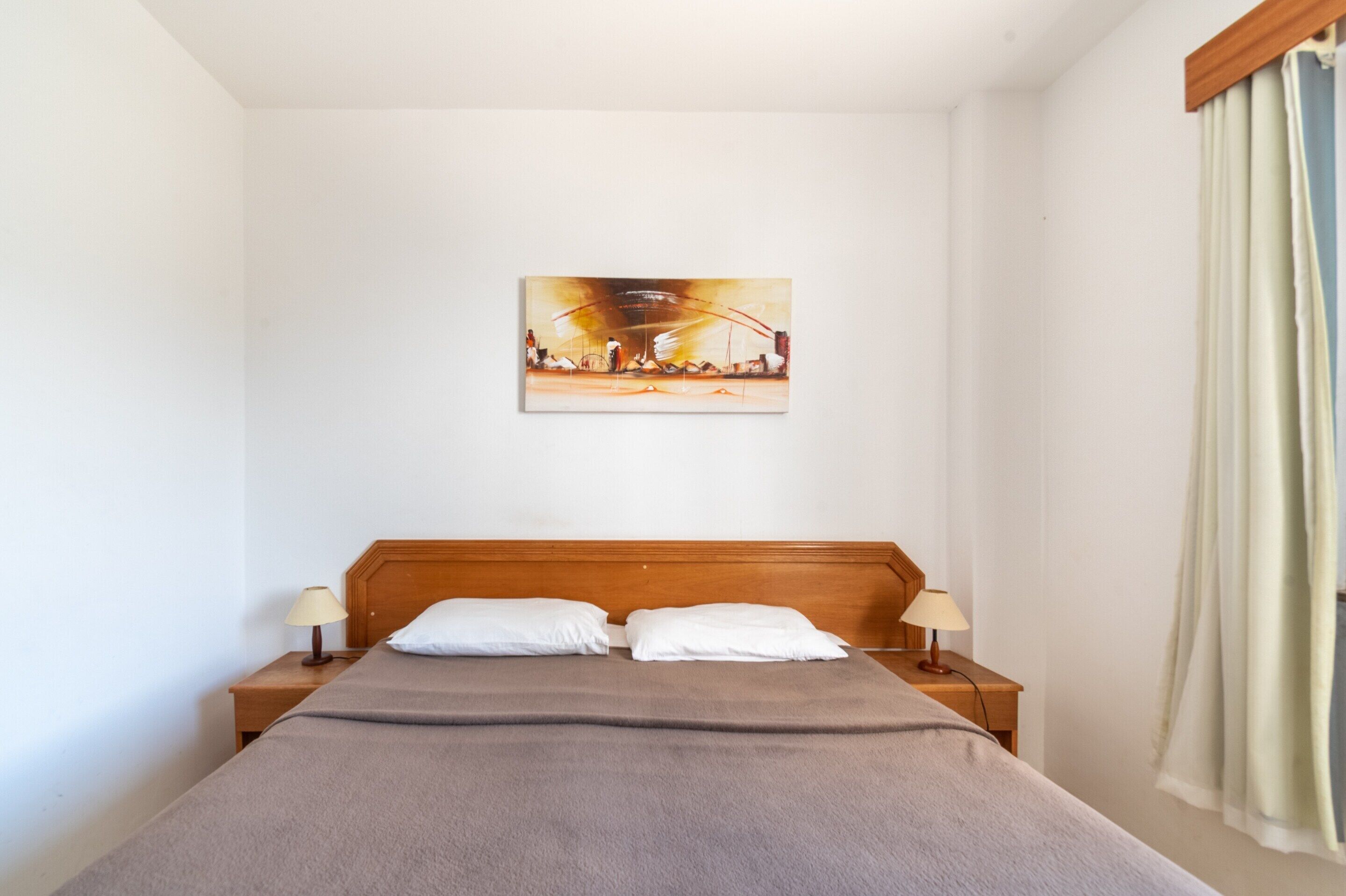 Habitación de hotel con cabecera de madera, cama con manta gris, dos almohadas blancas, dos lámparas de noche y un cuadro en la pared.