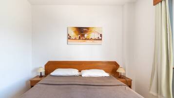 Habitación de hotel con cabecera de madera, cama con manta gris, dos almohadas blancas, dos lámparas de noche y un cuadro en la pared.