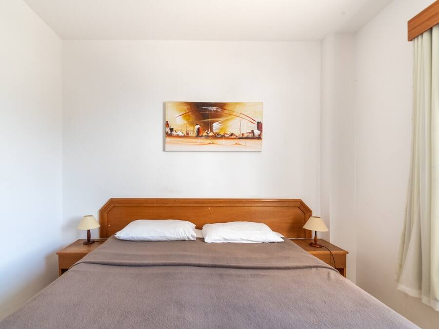 Habitación de hotel con cabecera de madera, cama con manta gris, dos almohadas blancas, dos lámparas de noche y un cuadro en la pared.