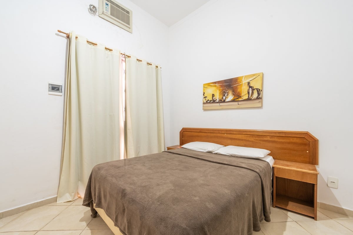 A bedroom with a bed, a wooden nightstand, a painting on the wall, and a window with curtains.