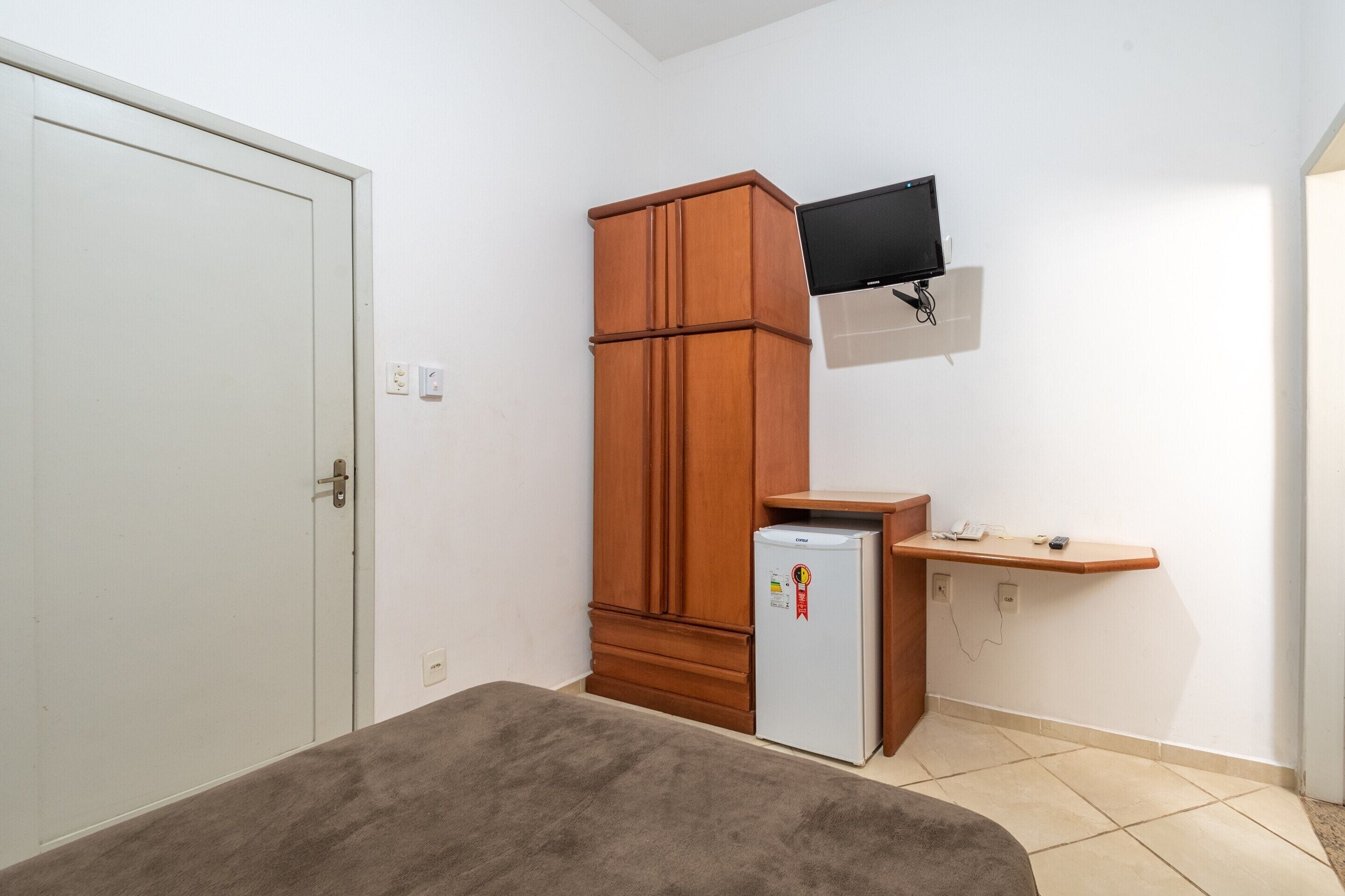 Ein Zimmer mit weißer Tür, einem Holzschrank, einem kleinen Kühlschrank, einem an der Wand montierten Fernseher und einem Holzregal mit Telefon und Fernbedienung.