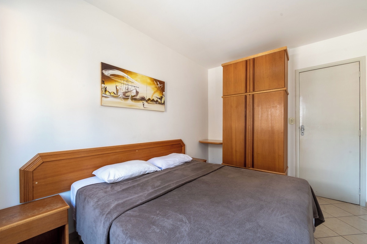 A bedroom with a bed, a wooden wardrobe, a nightstand, a painting on the wall, and a door.
