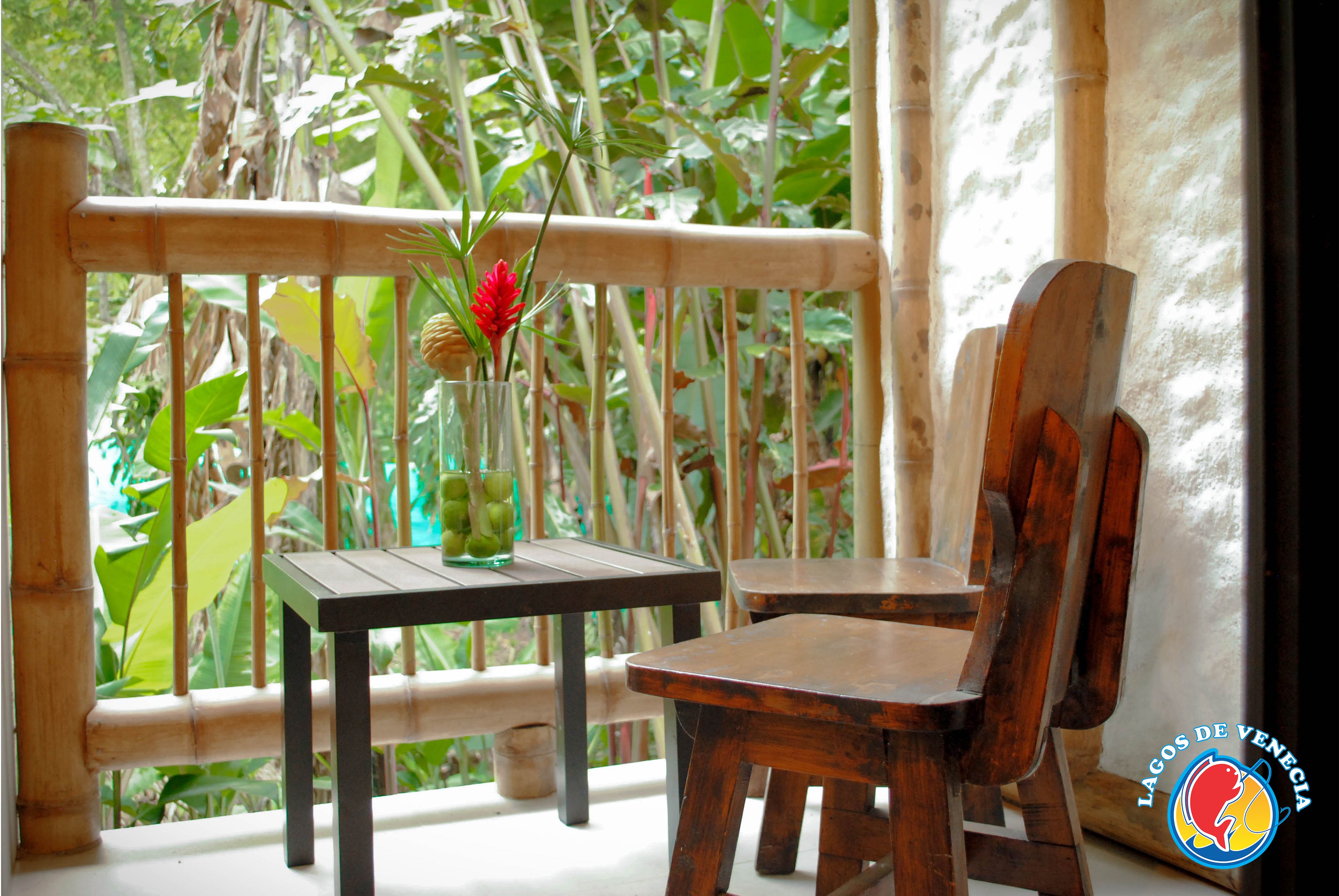 A wooden chair and table on a balcony with a bamboo railing, surrounded by lush greenery.