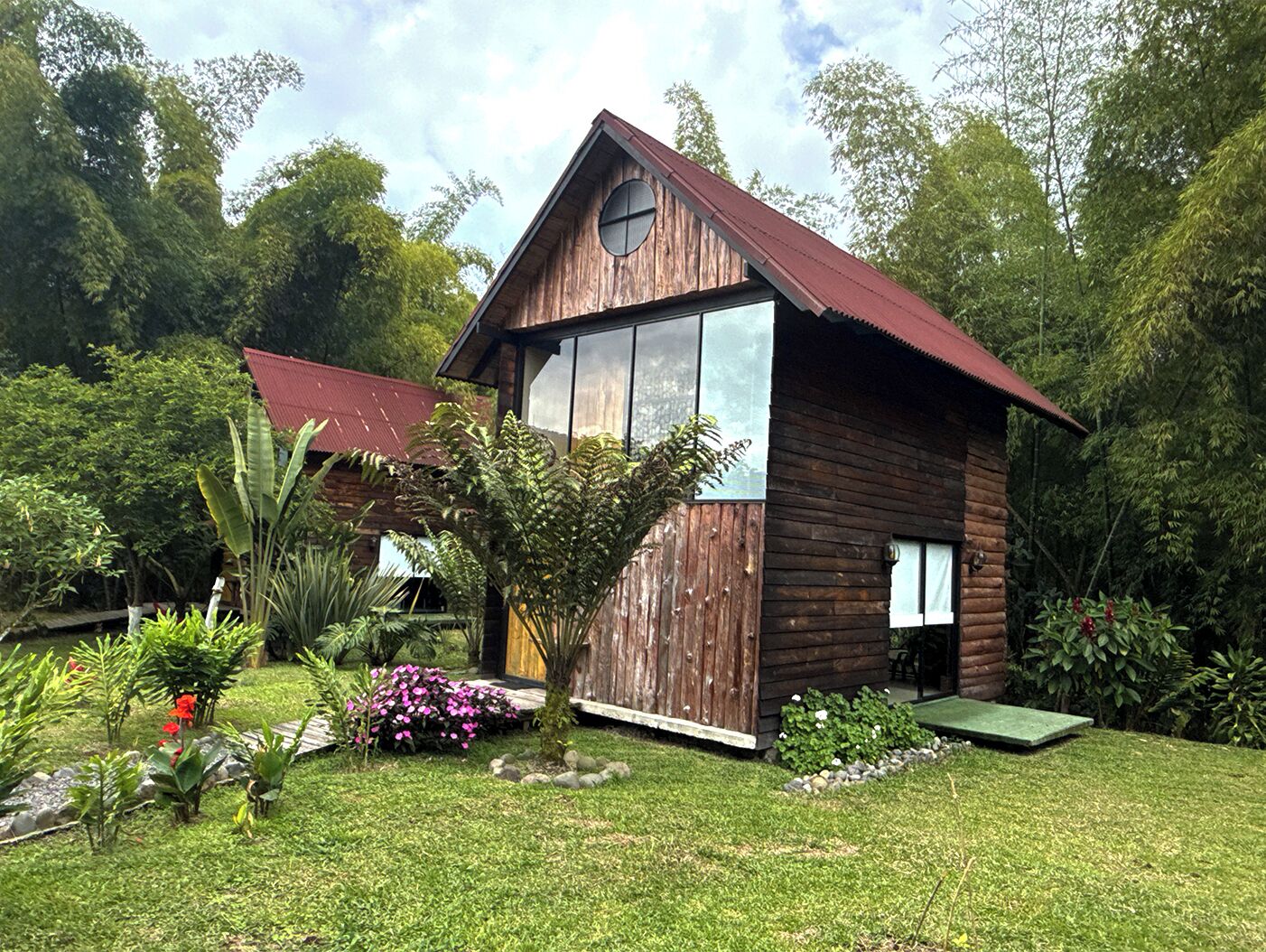 Una cabana de fusta amb una finestra gran i un teulat vermell, envoltada de vegetació exuberanta i un jardí ben cuidat.