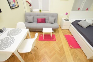 A compact room with a grey sofa, a small white table, a bed with a grey mattress, and a red rug. - Elegant Apartament Copernicus (Warsaw)