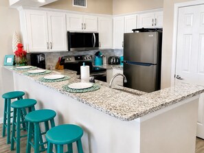 A kitchen with white cabinets, a stainless steel refrigerator, a microwave, and a stove. There are four teal stools and a countertop with a paper towel holder.