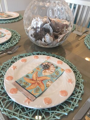 A table set with a glass bowl containing seashells, a decorative plate with a starfish design, and a woven placemat. - 3bdrm/3bath- BEACHFRONT AND 5 steps to TWO POOLS - sleeps 8! (Dauphin Island)