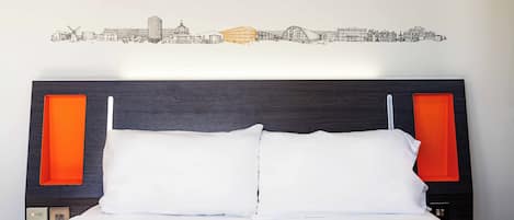 Una cama con ropa blanca, un cuadro de arte con un paisaje urbano y dos paneles naranjas en la cabecera.