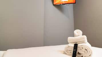 A hotel room with a bed, a TV mounted on the wall, and a stack of rolled towels on the bed.