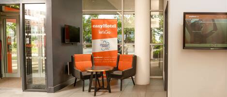 Zona de recepción con un cartel de easyHotel, una pantalla de televisión y un área de descanso con una mesa.