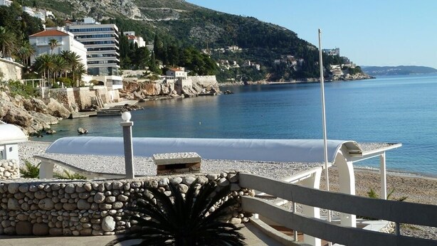 A coastal view with a stone wall, a palm tree, and buildings along the shoreline.