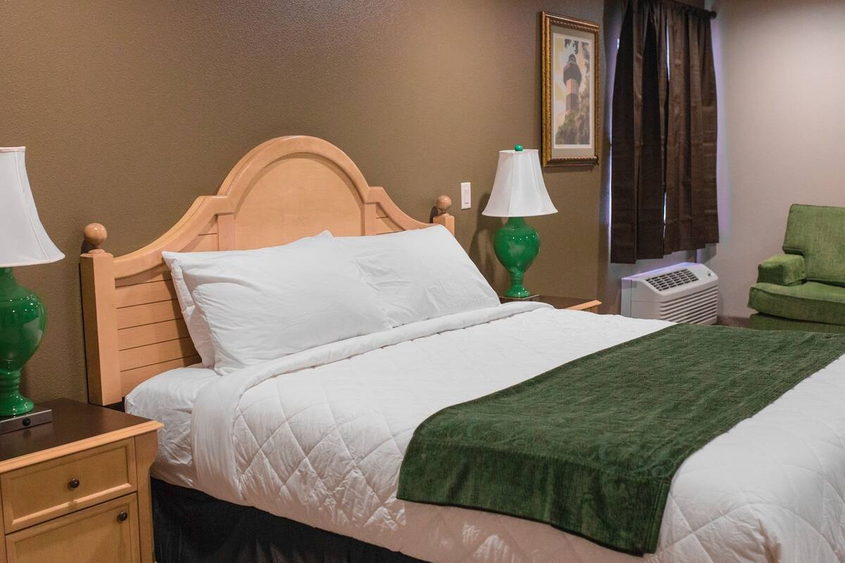 A neatly made bed with a wooden headboard, two bedside lamps, a green throw blanket, a green chair, and a framed picture on the wall.