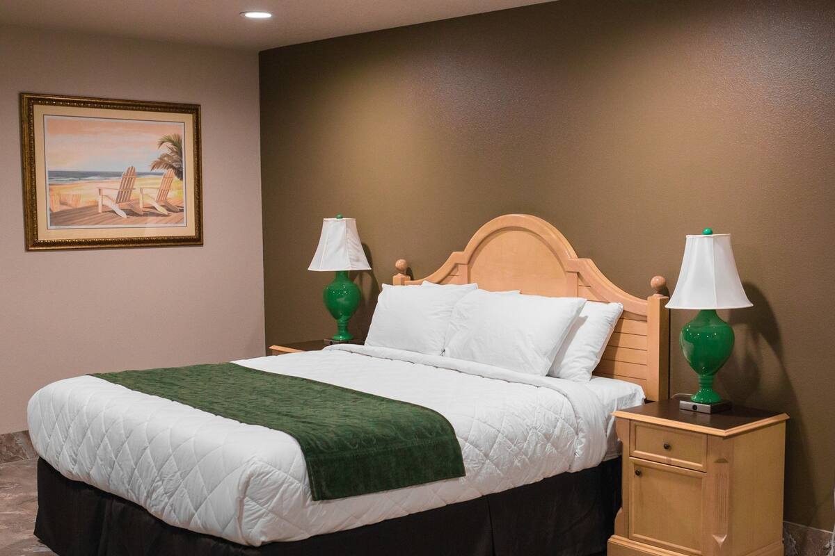 A neatly made bed with a green throw, two bedside lamps, a wooden nightstand, and a painting of a beach scene on the wall.
