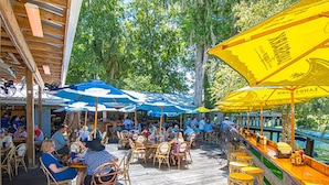 An outdoor dining area with wooden tables and chairs, covered by blue and yellow umbrellas, overlooking a body of water. - The Gator Den Motel (Dunnellon)