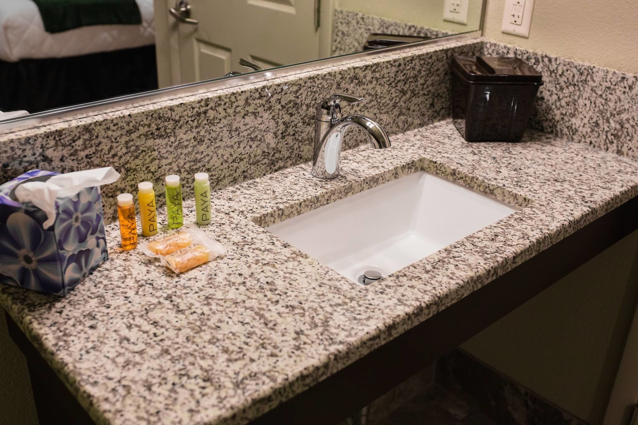 A granite countertop with a white sink and a mirror above it. There are toiletries on the counter, including bottles and a tissue box.