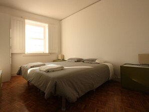 Quarto com duas camas, soalho de madeira, janela com portadas e uma mesa de cabeceira com abajur, além de uma cômoda verde.
