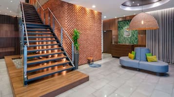 Un moderno vestíbulo de hotel con una escalera de madera, un mostrador de recepción, una zona de estar y una lámpara colgante grande.