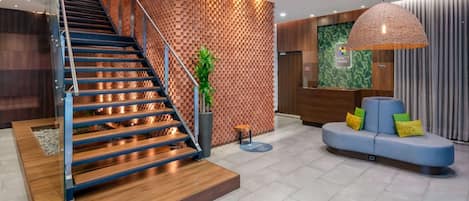 A modern hotel lobby with a wooden staircase, a reception desk, a seating area, and a large pendant light.
