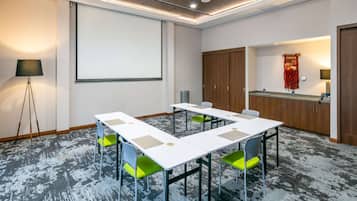 Een vergaderruimte met een witte tafel, groene stoelen, een scherm voor een beamer en een kitchenette.