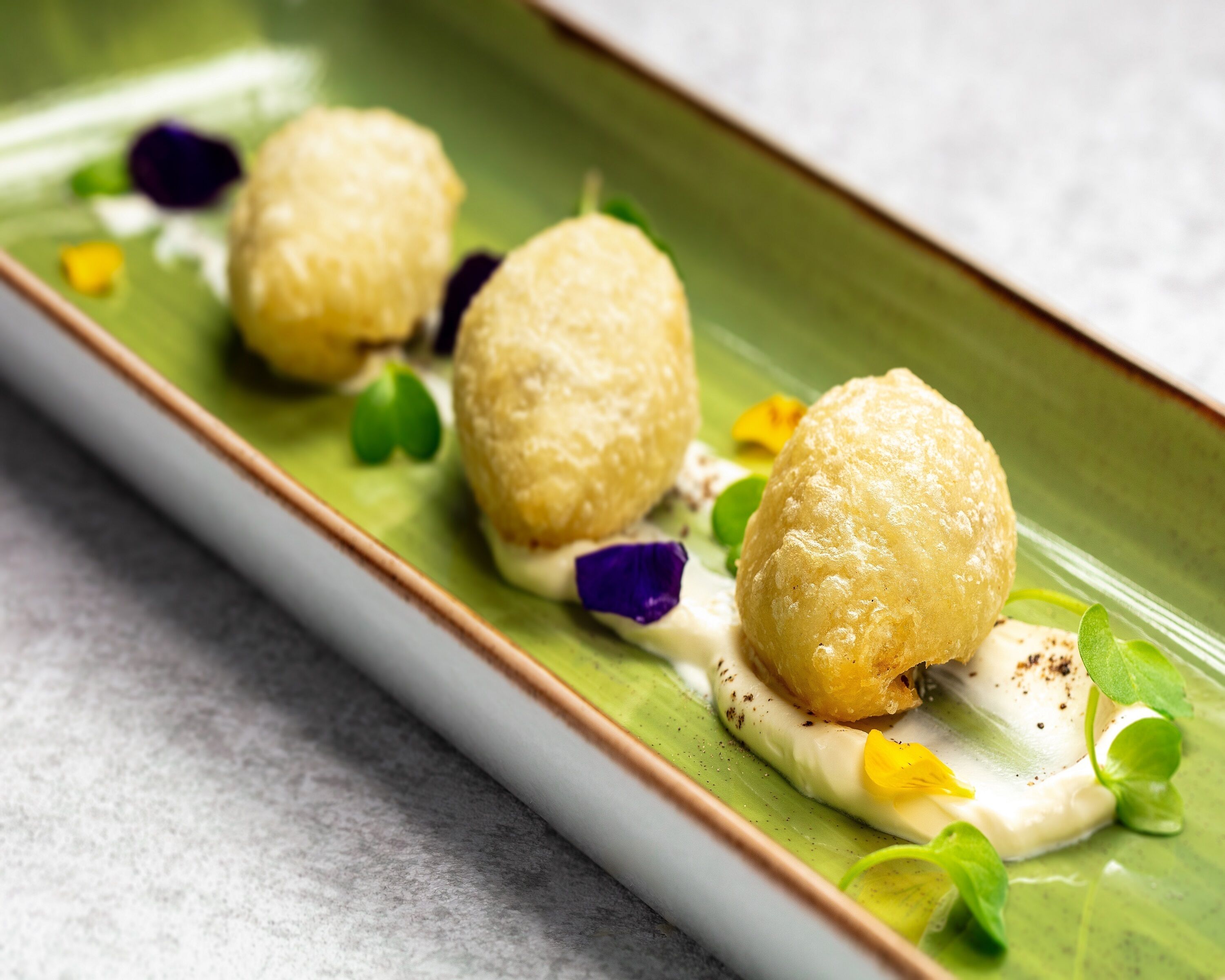 Un plato rectangular con tres elementos redondos, de color marrón dorado, posiblemente comida, acompañado de hojas verdes y flores moradas.